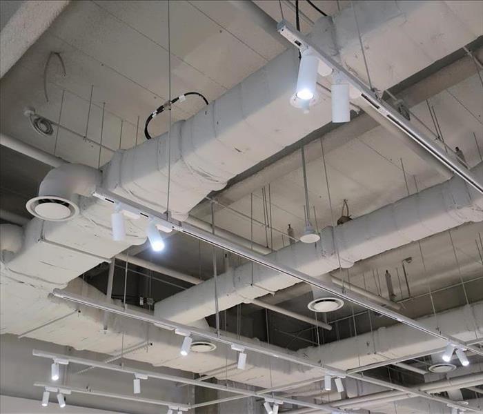 The ceiling of a commercial facility showing exposed air ducts with circular vents that and hanging light fixtures 